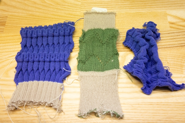 Three samples of machine-knit material lay on a table - two purples on either end and a green one in the middle.