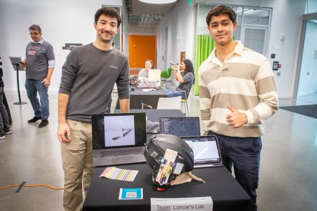 Two Harvard students behind a black skiing helmet modified with electronic sensors and a laptop display