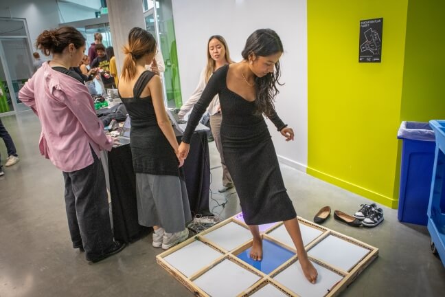 A female Harvard student wearing a black dress stands over a wooden board with light-up panels in front of a group