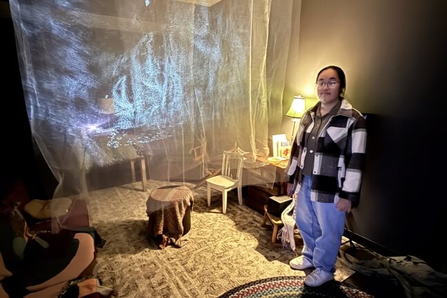 A female Harvard Student in an art installation that resembles a living room with swirling blue images projected onto tulle sheets