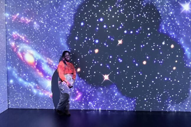 A female Harvard student standing next to a digitally projected image of stars, with the silhouette of a Black woman's head superimposed