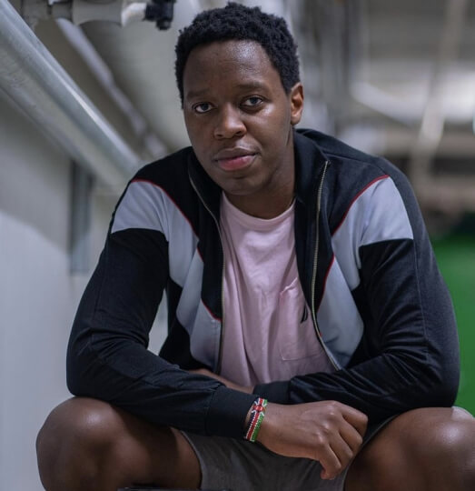 A young man in a light pink shirt and dark gray jacket sits and leans forward, looking directly at the camera.