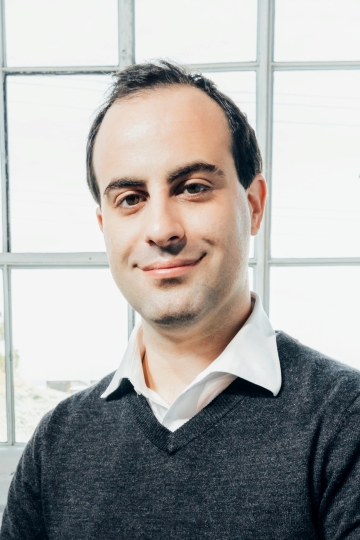 A man in a white button down and dark gray sweater stands in front of a brightly lit window. 