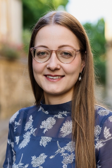 A woman with glasses and a printed blue shirt smiles in front of a blurred outdoor background.