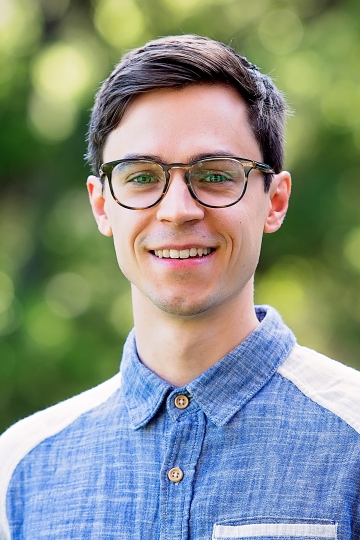 A man in a denim-textured blue shirt and glasses smiles in front of a blurred green background.