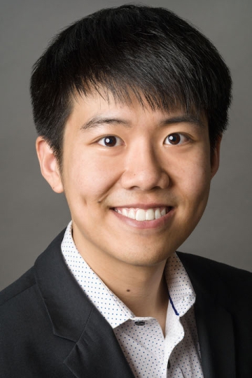 A man in a white polka dot button down and black blazer smiles in front of a plain gray background.