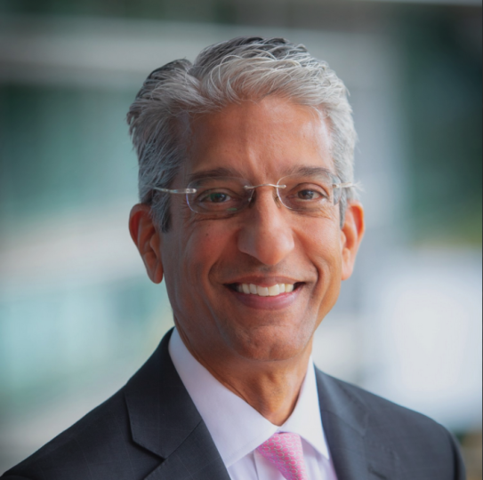 A man with white/gray hair and frameless glasses smiles in front of a blurry background. 