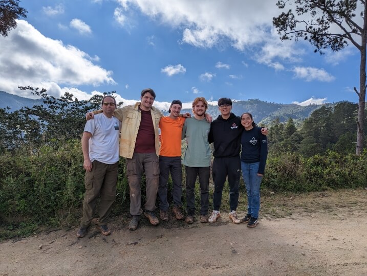 Six people standing on a dirt road under a partially cloudy sky with mountains in the background