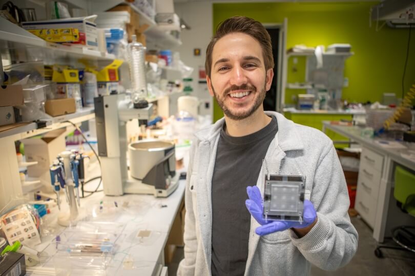 Harvard SEAS graduate student Gino Domel wearing a white lab coat next to a table of laboratory equipment