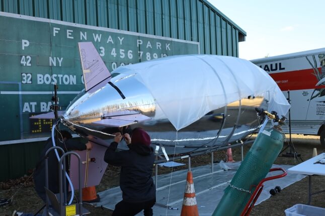 A large silver-colored blimp being filled with helium by Harvard SEAS students at Crow Island Airpark in Stow, Mass.